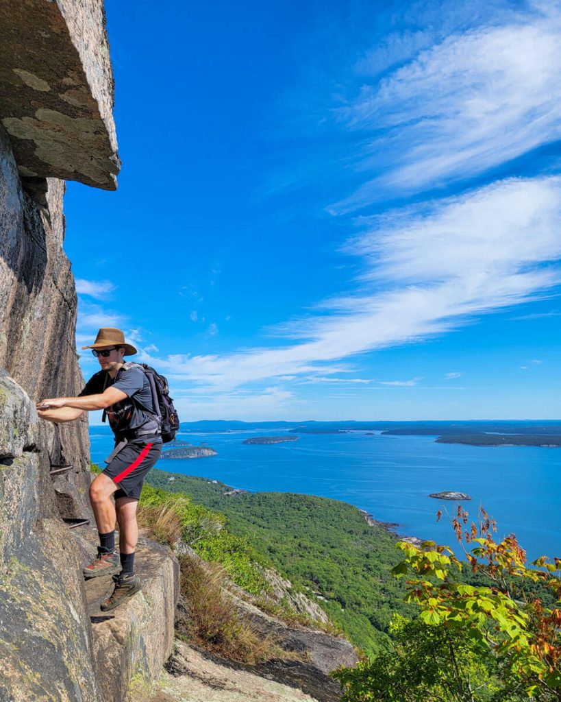 Hiker climbs on rungs and ladders of Precipice Trail in Acadia National Park in Maine with ocean in the background on a sunny day. 